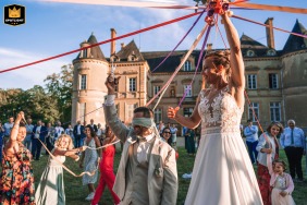 No cenário romântico do Castelo de Beaulon, Charente-Maritime, um noivo vendado tenta de forma divertida cortar a fita do buquê de sua noiva, capturando a atmosfera alegre e amorosa da celebração do casamento.